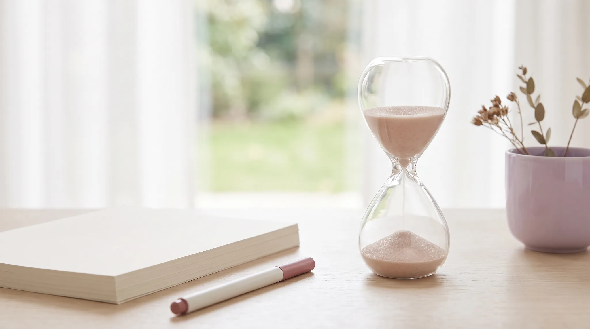 Sanduhr mit hellem Sand neben einem ordentlichen Papierstapel und einem Stift auf einem hellen Schreibtisch.